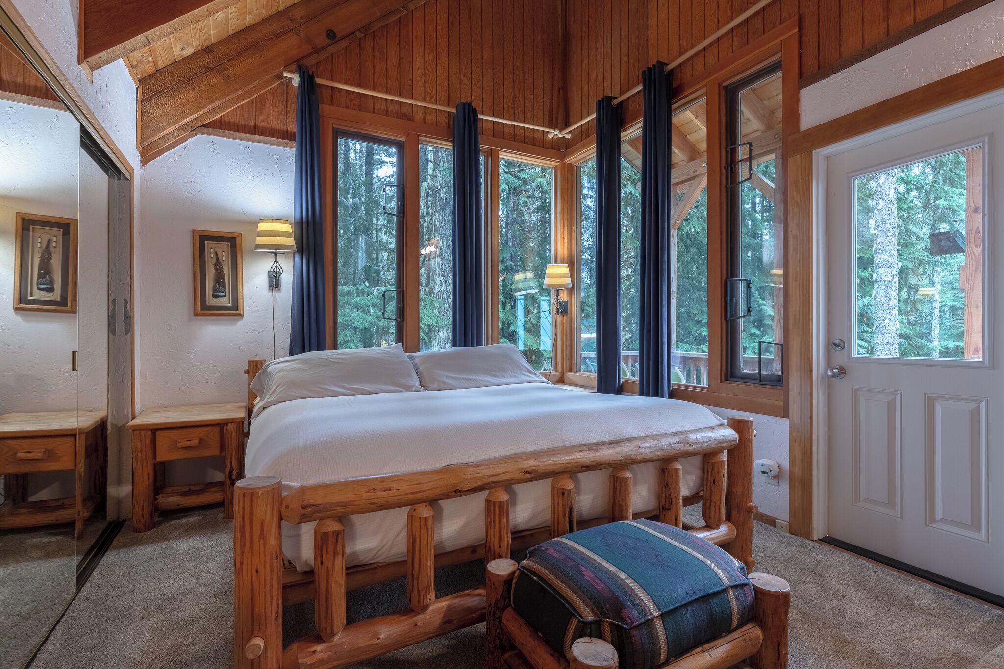 A wooden-framed bed with a bench, two bedside tables, and a door with a view of trees.