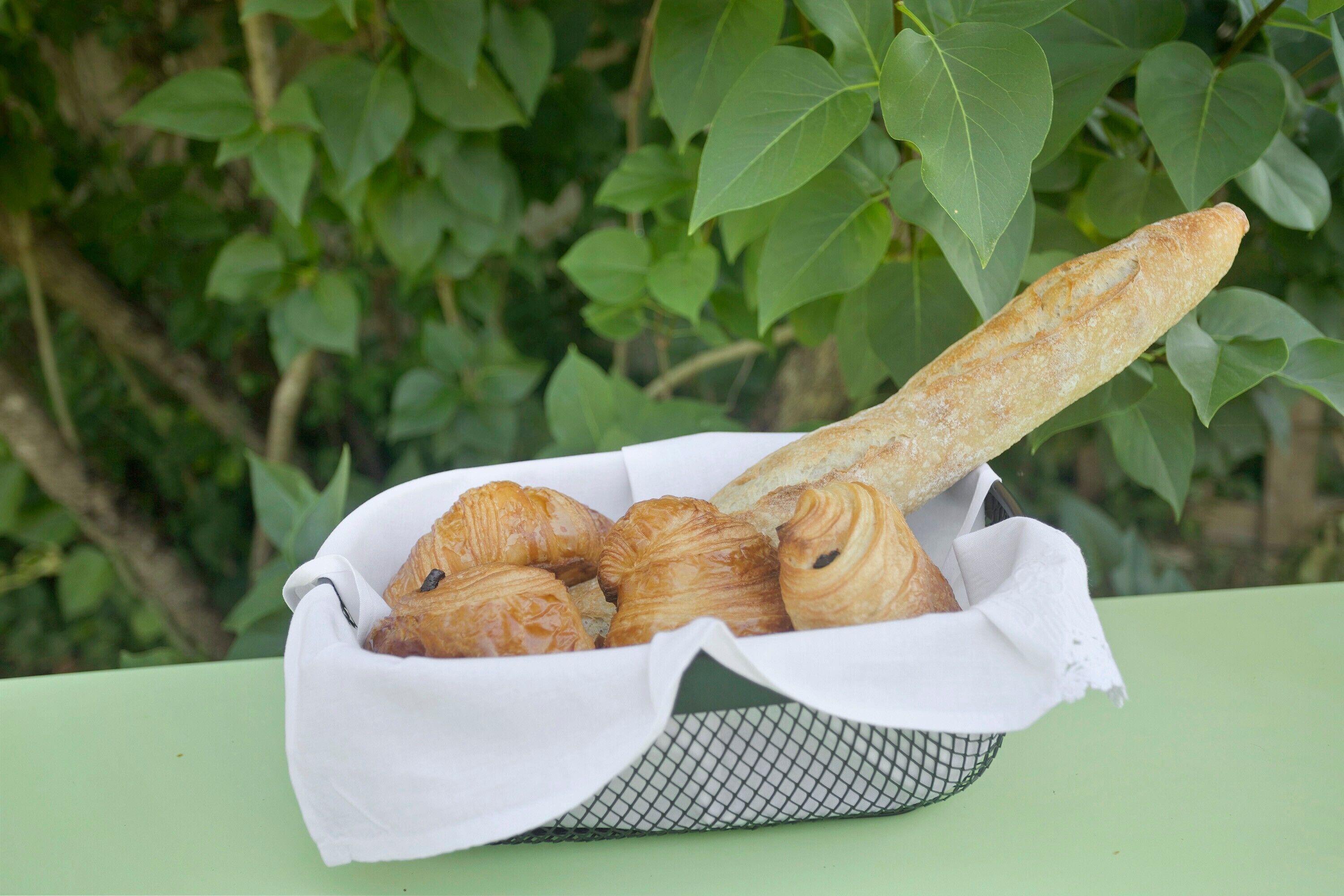 A basket of croissants and a baguette on a table with a green background.