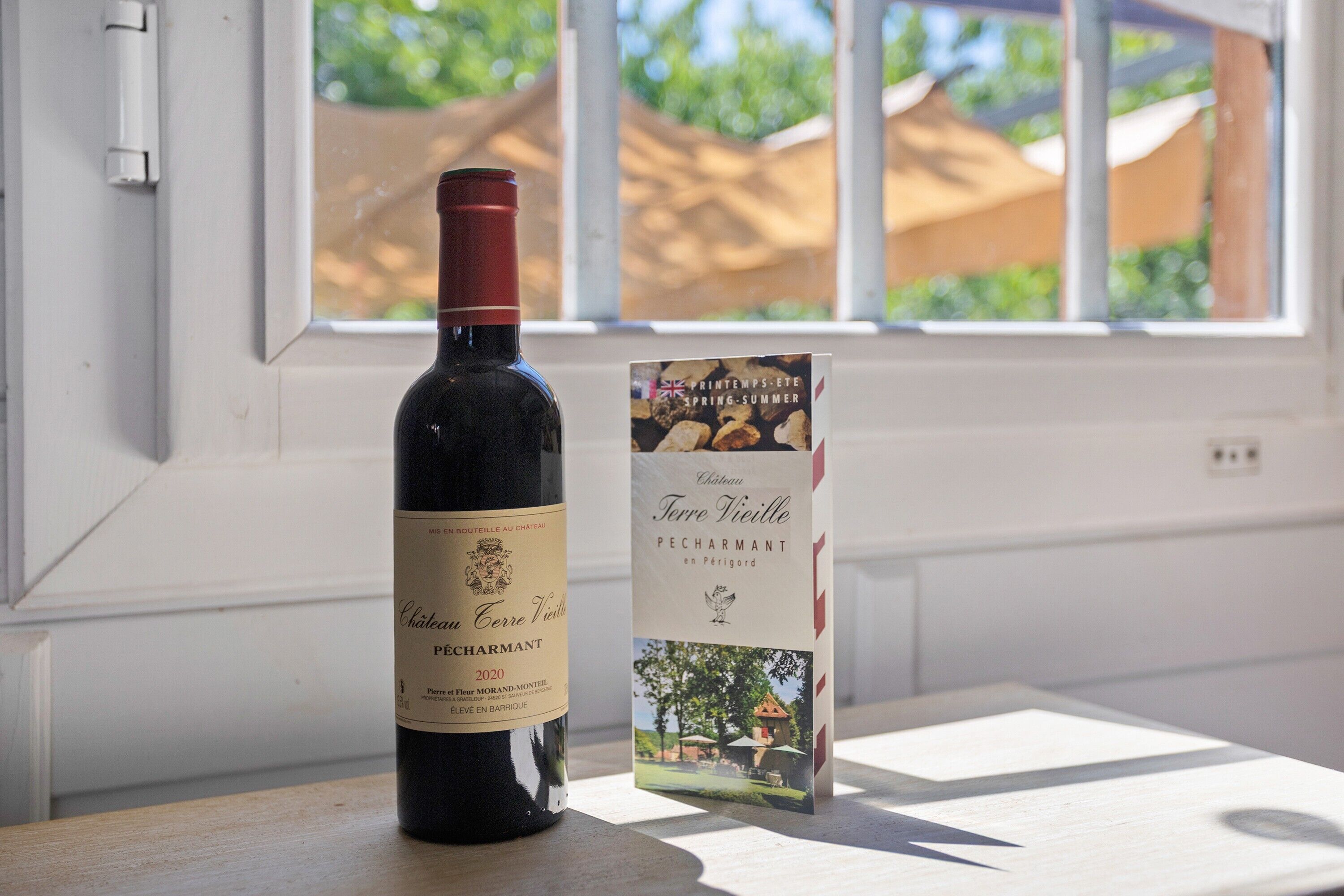 Une bouteille de vin Château Terre Vieille Pecharmant, millésime 2020, et son étiquette, posées sur une table près d’une fenêtre.