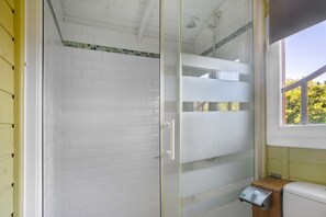 Un baño con cabina de ducha de vidrio, paredes cubiertas de azulejos blancos, una ventana y un taburete de madera.