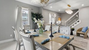 Un comedor con una mesa grande, sillas blancas y un candelabro. Se ve una escalera al fondo.