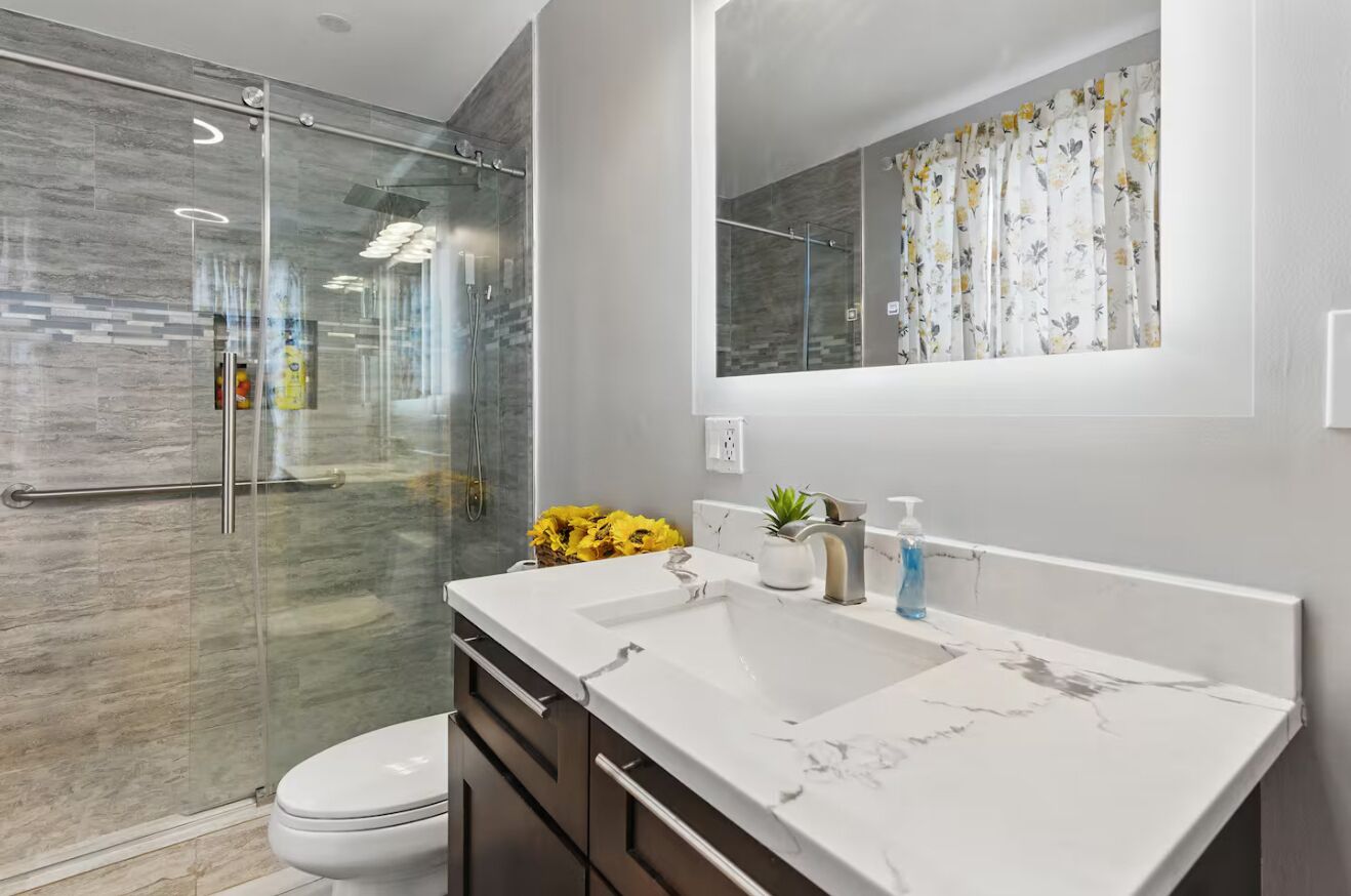 Un baño moderno con ducha envuelta en vidrio, lavamanos blanco y encimera de mármol, y un espejo grande.