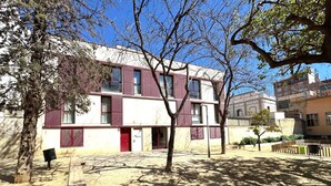 Um edifício de vários andares com fachada vermelha e branca, entrada visível e um pátio pavimentado com árvores.