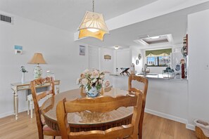 Un coin repas avec une table ronde, des chaises, un vase de fleurs, une lampe et, au fond, une cuisine.