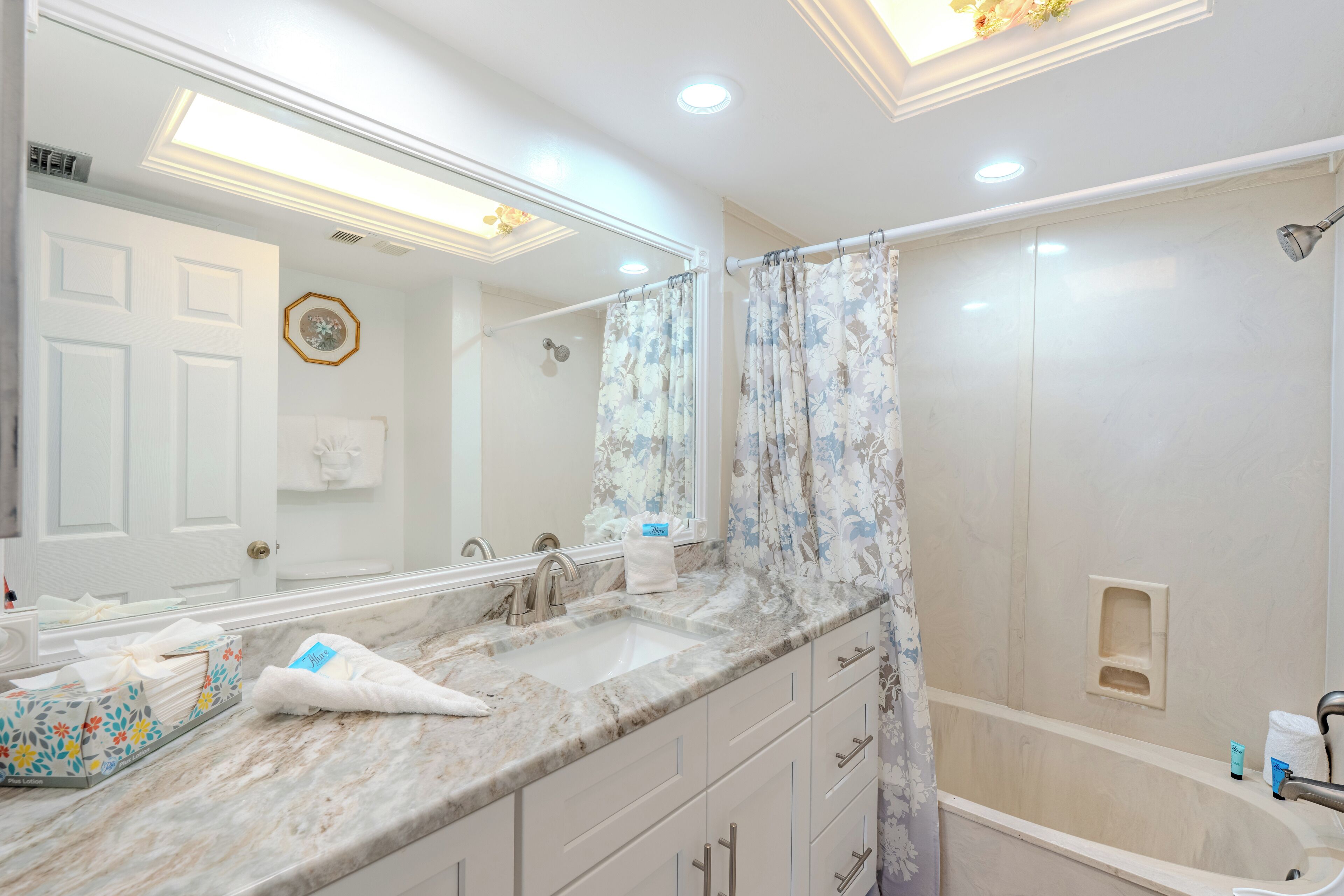 Un baño con encimera de mármol, dos lavamanos, un espejo grande, ducha con cortina estampada y un perchero.