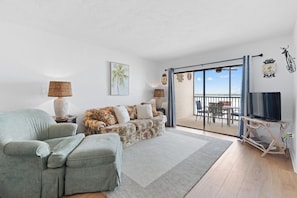 A living room with a sofa, a chair, a television on a stand, and a balcony with a view.
