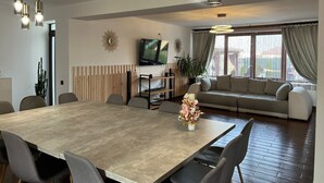 A modern living room with a dining area, featuring a large wooden table, grey upholstered chairs, a sofa, and a TV mounted on the wall. - Mik Chalet. Pet friendly (Săcele)