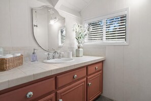A bathroom with a white vanity, a large mirror, a window with blinds, and a vase with flowers. - Walk to the lake Gorgeous lake house (Big Bear Lake)