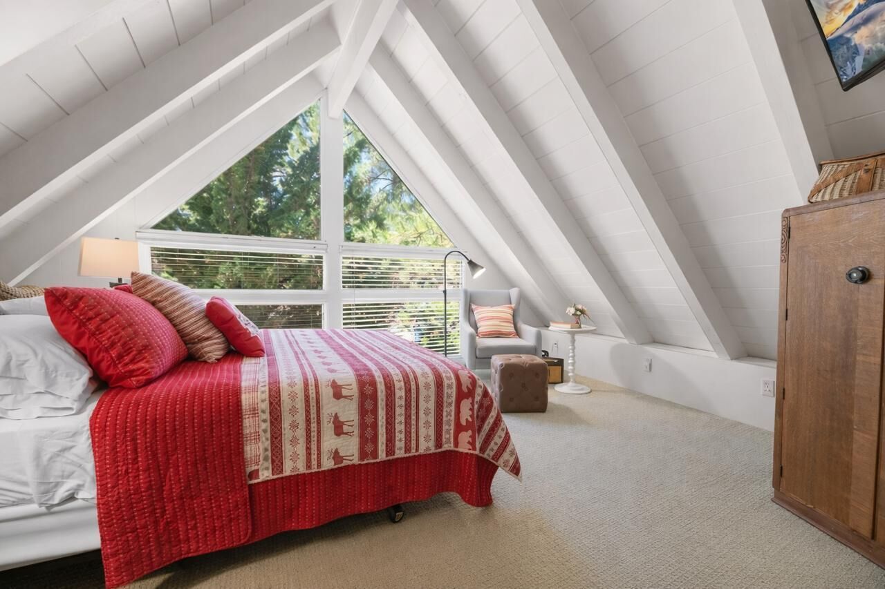 A bedroom with a sloped ceiling, a large window, a bed with a red blanket, a chair, and a wooden cabinet.