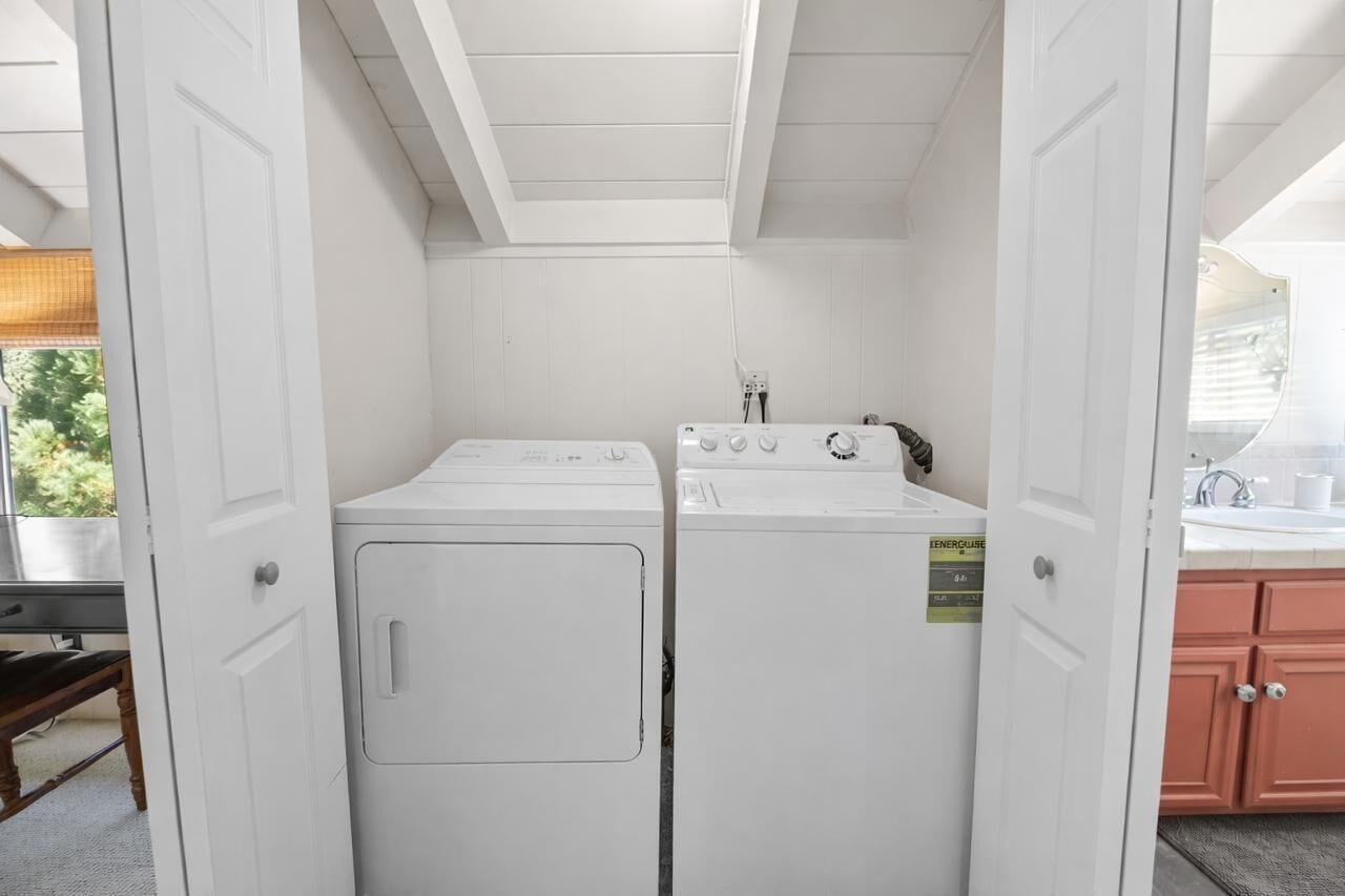 A laundry room with two white washers and dryers, a wooden bench, and a window with a view of greenery.