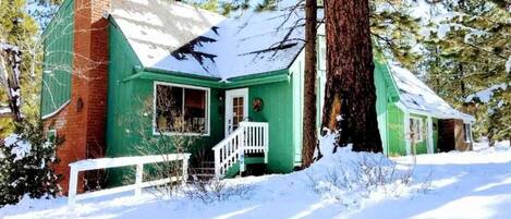 Une cabane verte avec un porche blanc, entourée de neige et d’arbres.