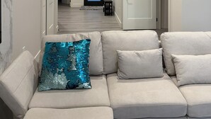 Ett modernt vardagsrum med en beige hörnsoffa, en dekorativ turkosfärgad kudde och en hall som leder till en dörr med en blått upplyst panel.