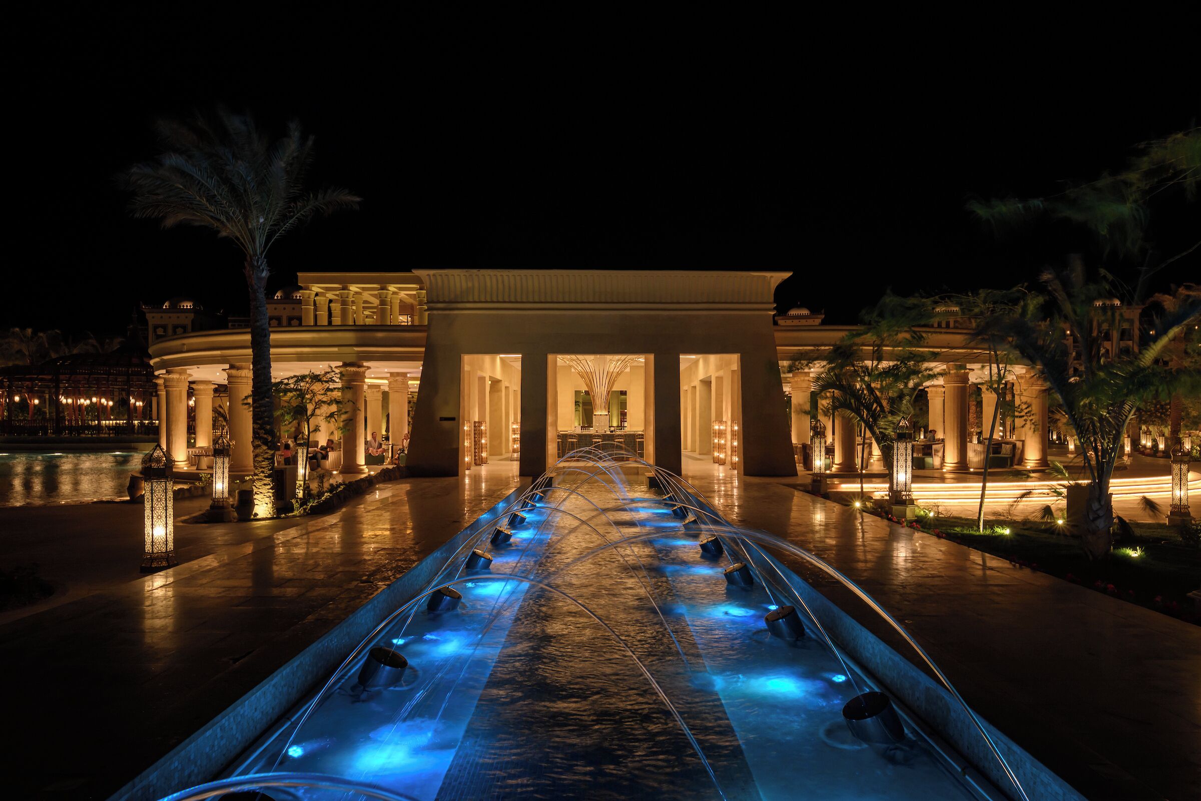 A nighttime view of a grand hotel entrance with a water feature, palm trees, and classical architecture.