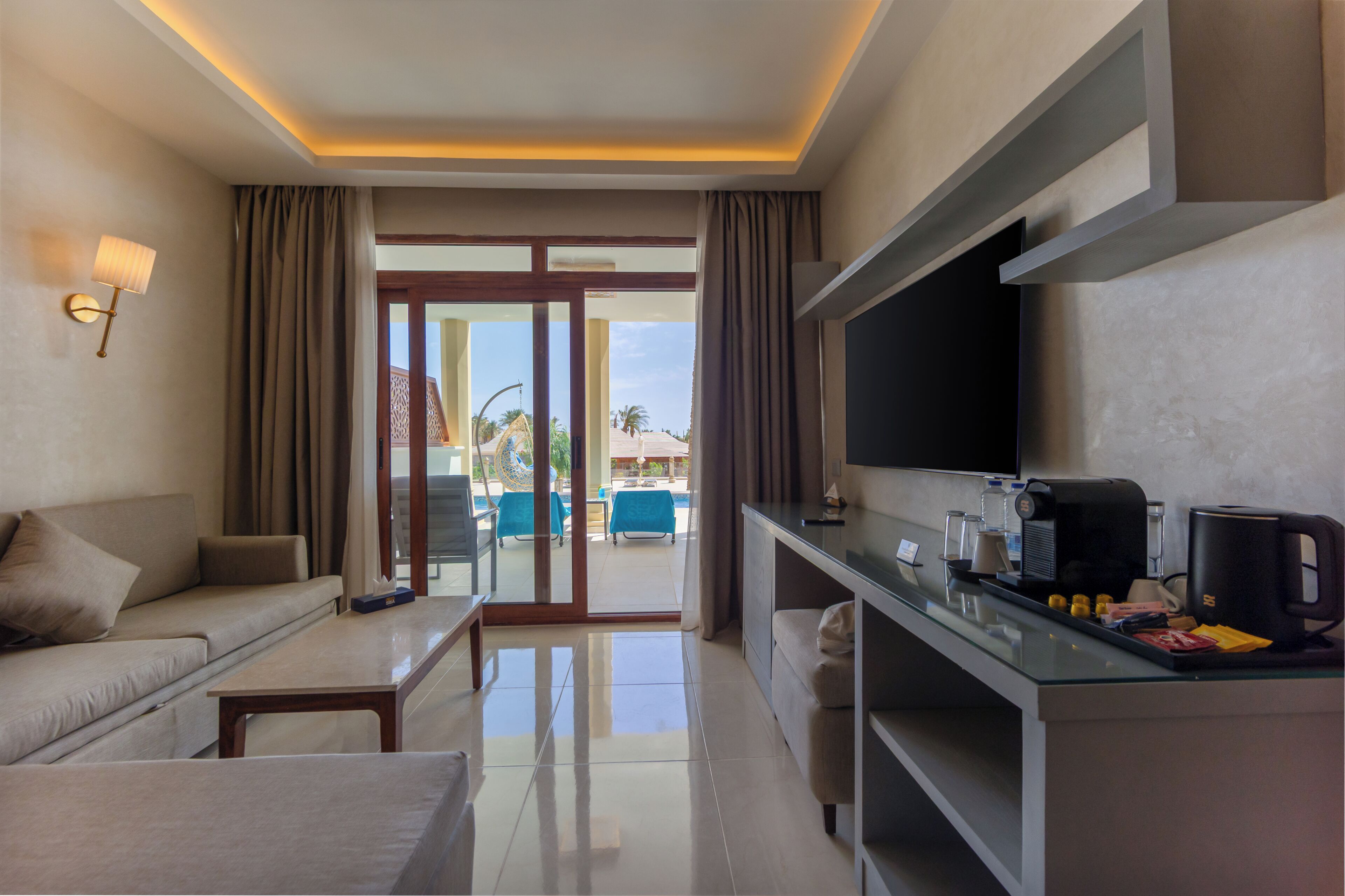 A hotel room with a sofa, a coffee table, a TV, a coffee maker, and a view of the pool and palm trees.