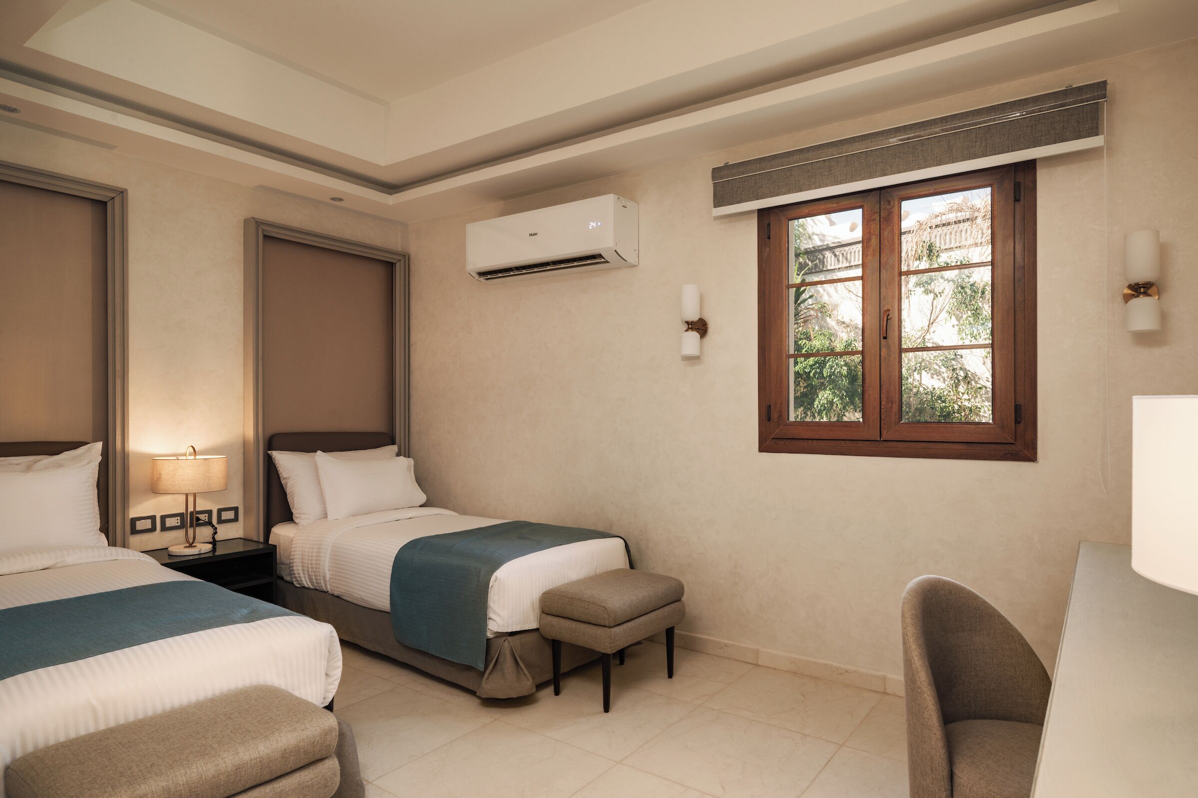 A hotel room with two beds, a desk, a chair, an air conditioning unit, a window with a view, and a lamp on a bedside table.