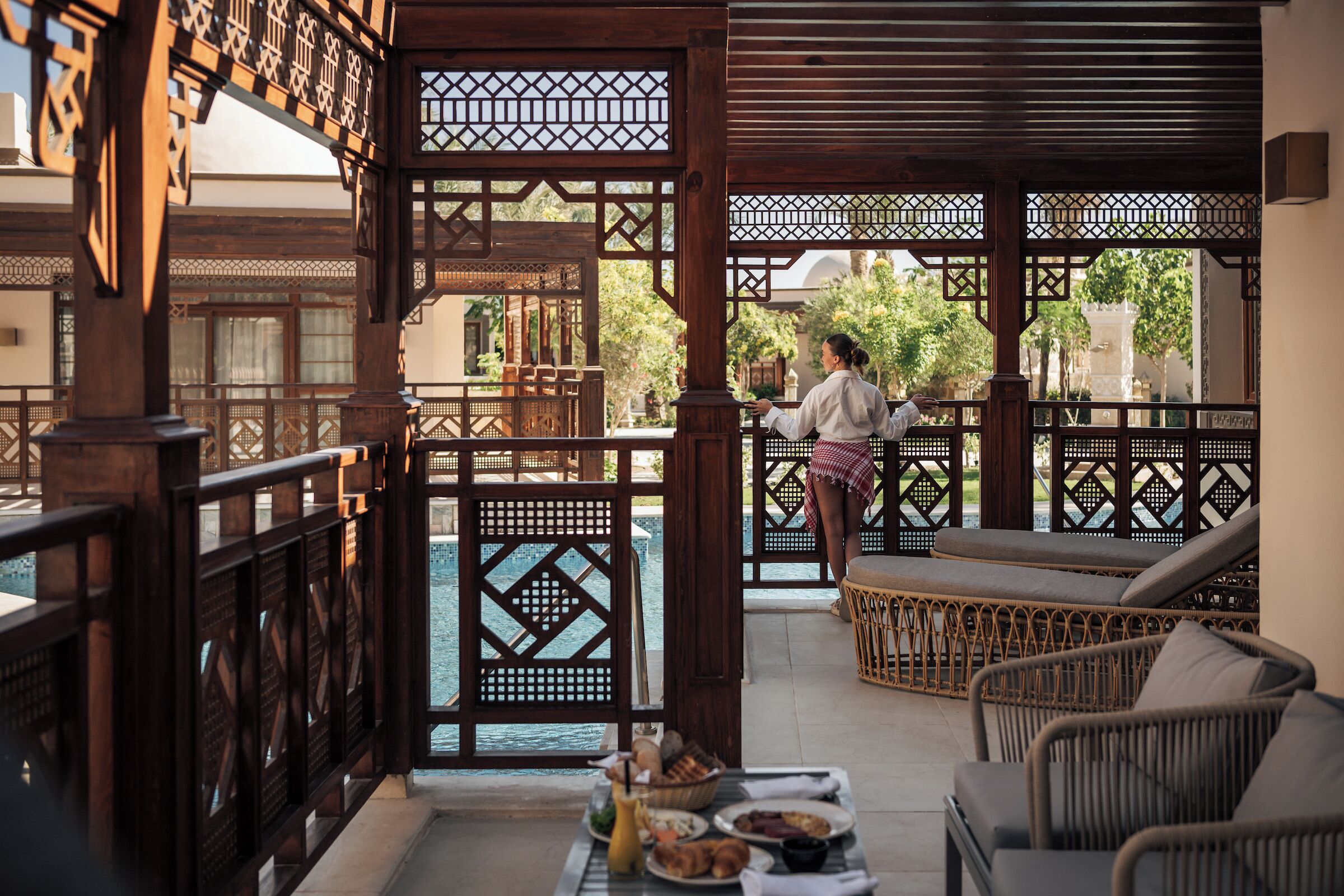 Un balcon traditionnel en bois offrant une vue sur la piscine et le jardin, avec des meubles en osier et une table où sont disposés des éléments pour le petit-déjeuner.