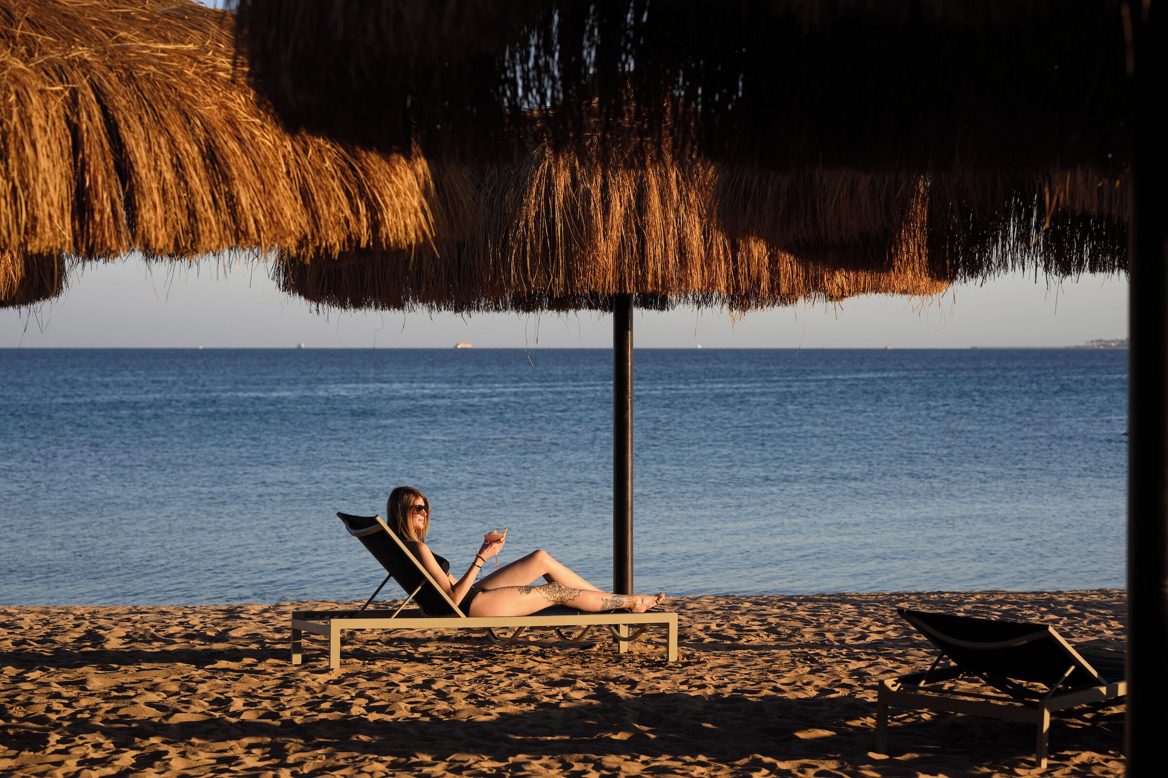 A beach scene with a person relaxing on a lounge chair under a thatched umbrella, a second lounge chair, and the ocean in the background.