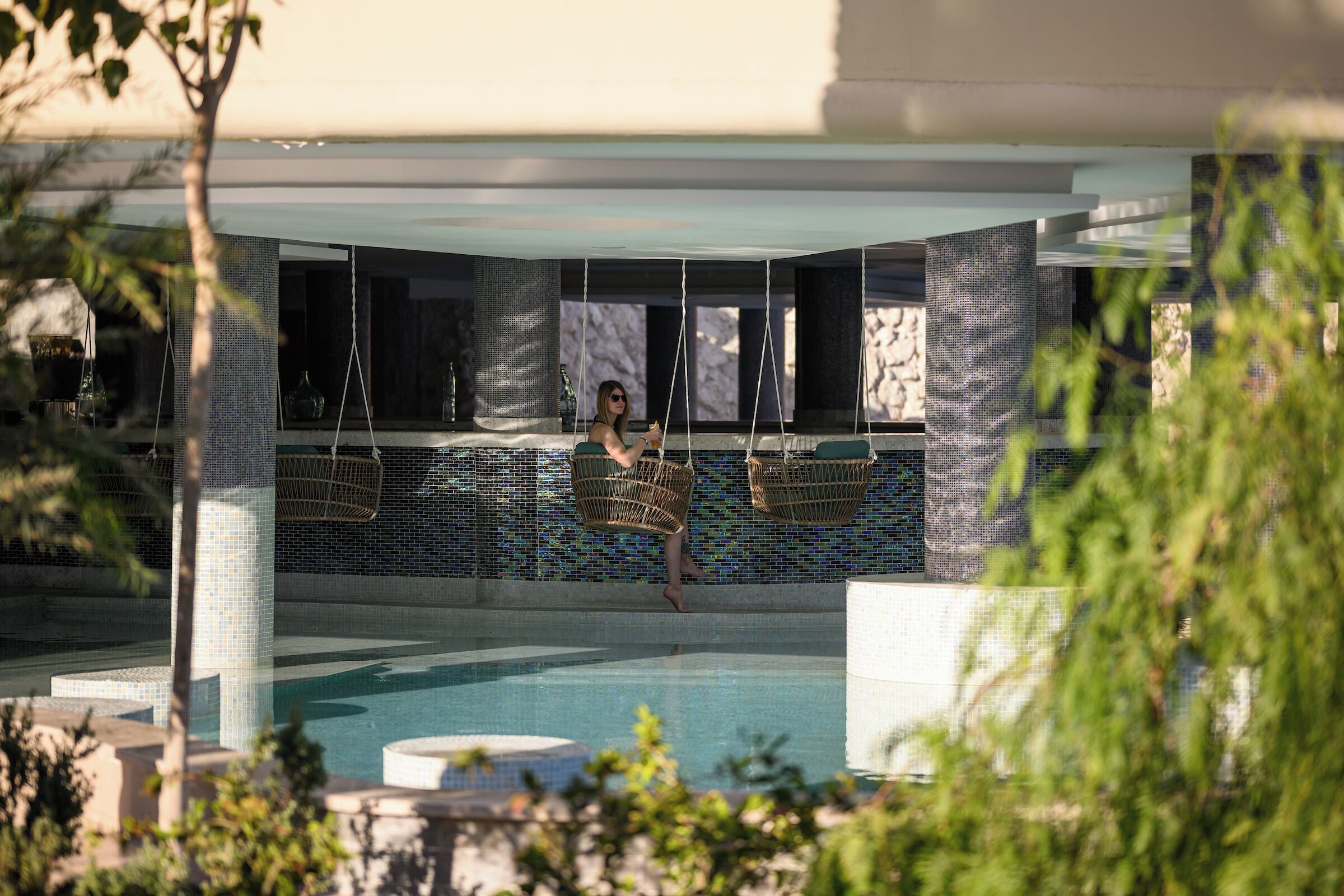 Zona de piscina con cestas colgantes, pared de azulejos y una persona relajándose junto a la piscina.
