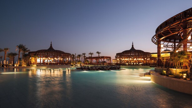 A resort pool area with lounge chairs, palm trees, and illuminated structures at dusk. - Al Kasr Sahl Hasheesh-Ultra All-Inclusive (Sahl Hasheeh)