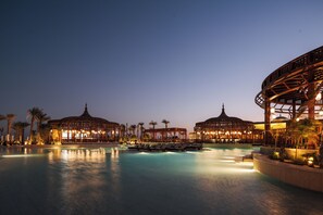 Zona de piscina en un complejo turístico con sillones de descanso, palmeras y estructuras iluminadas al anochecer.