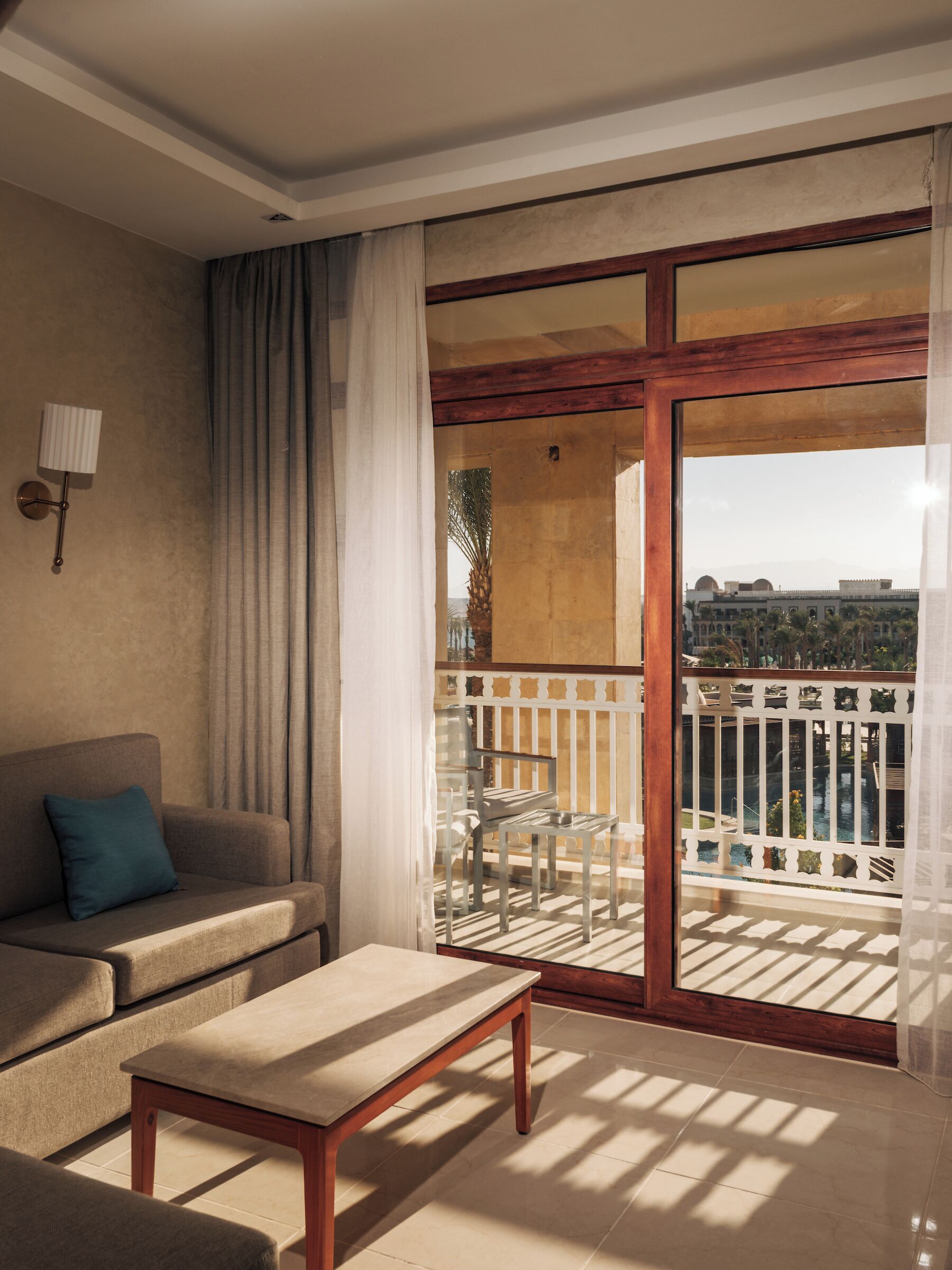 A room with a sofa, a table, a balcony with a railing, and a view of a building and palm trees.
