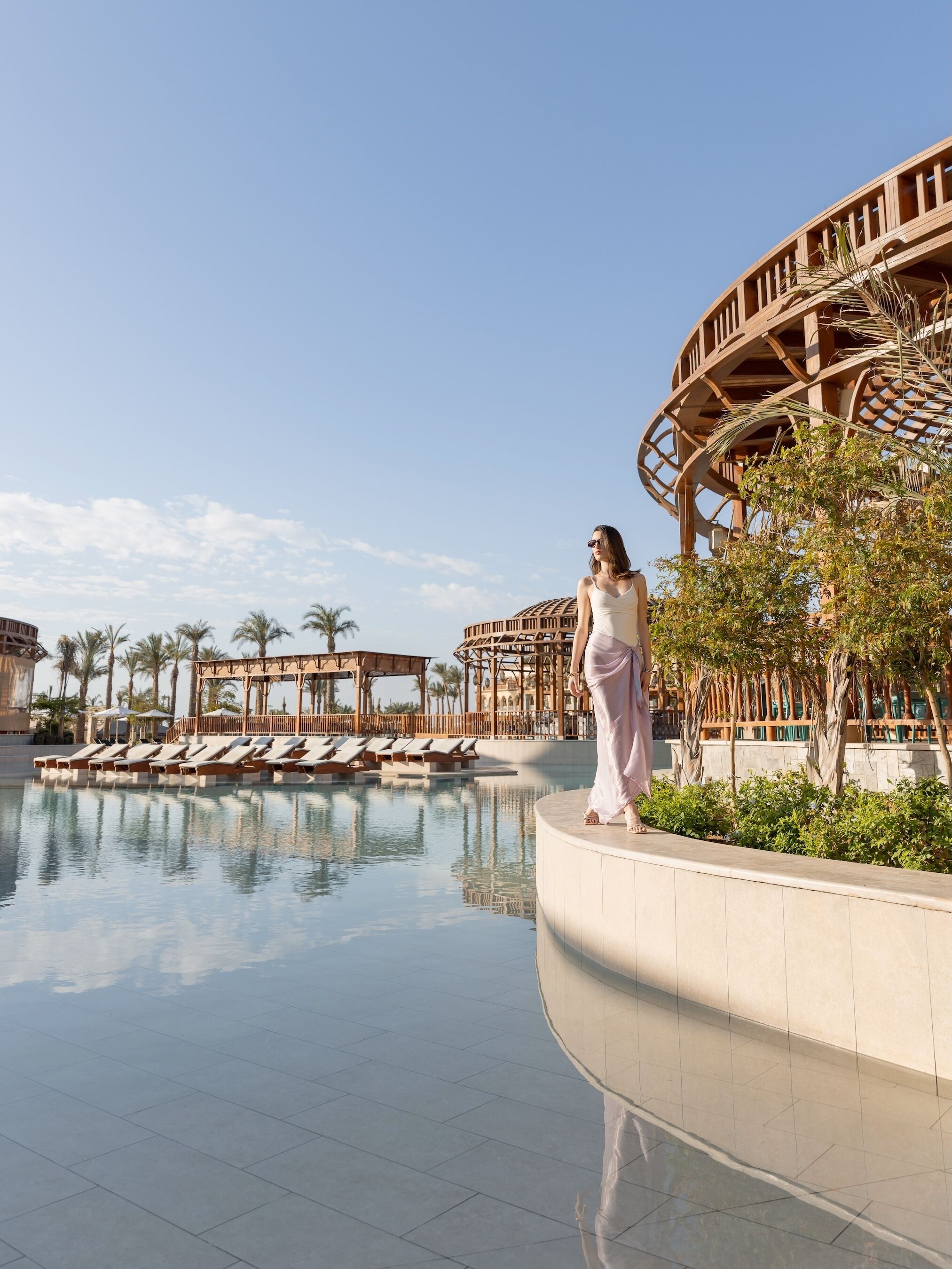 A poolside area with lounge chairs, a wooden pergola, and a clear sky.