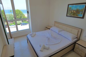A bedroom with a bed, a nightstand, a drawer, and a window with a view of the beach and cars. - Almare Apartments (Orikum)
