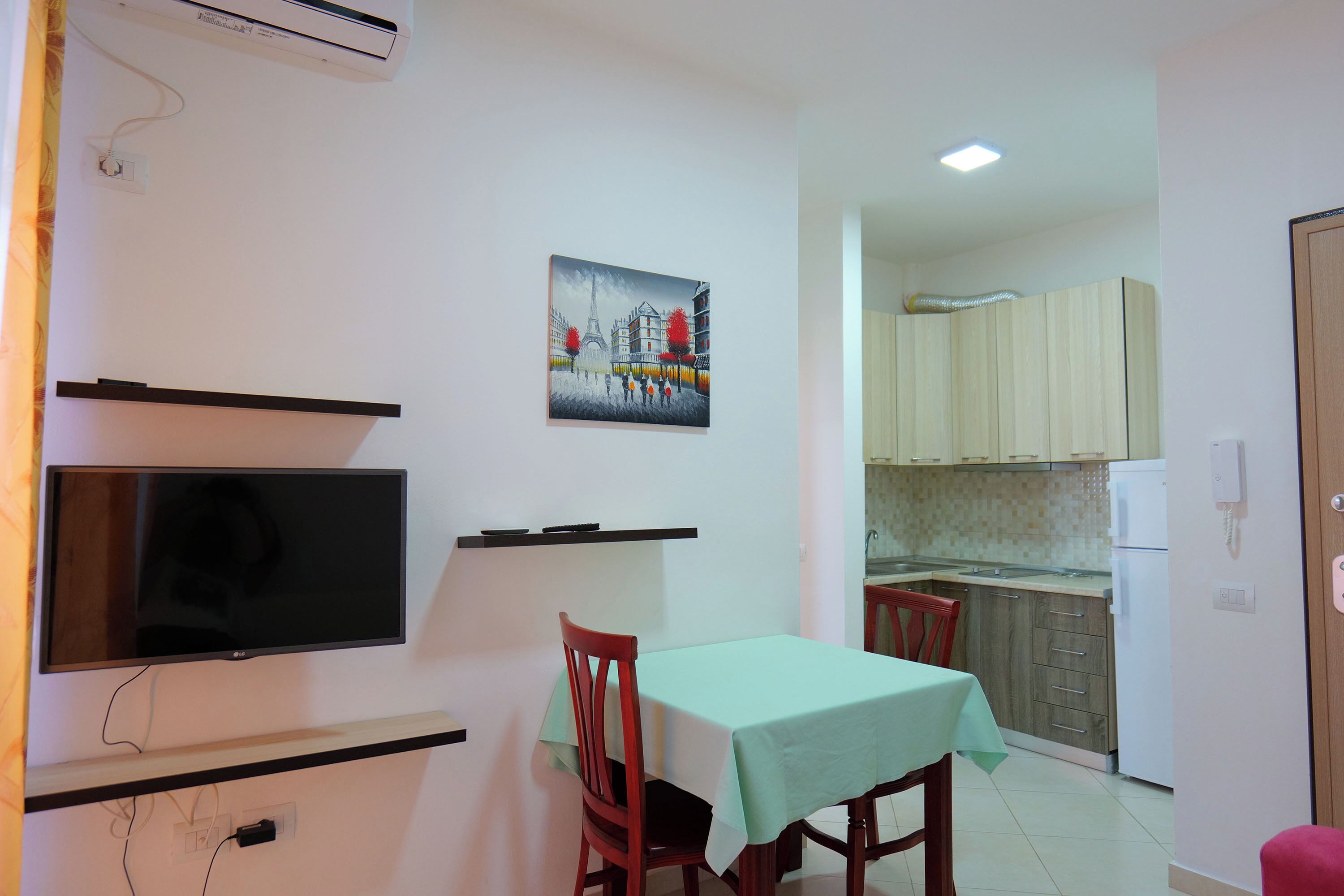 A dining area with a table, chairs, a TV, and a kitchen in the background.