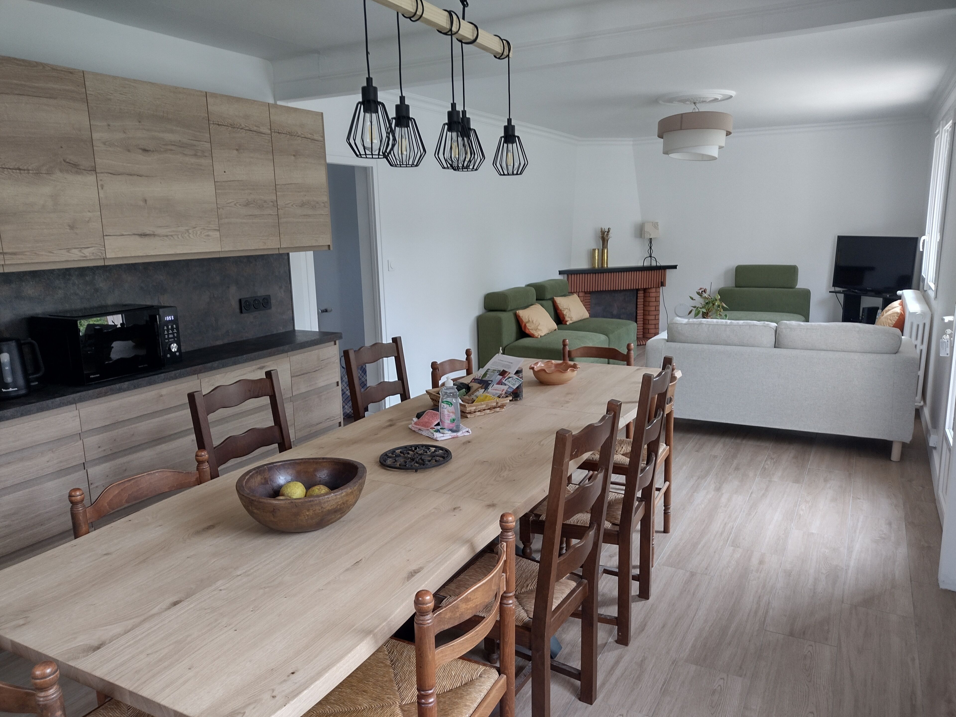Une cuisine moderne avec des meubles en bois, une table à manger avec des chaises, un four à micro-ondes et des luminaires suspendus.