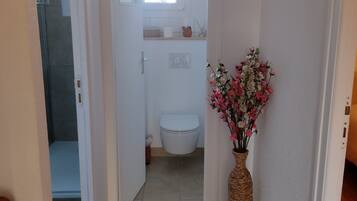 Een smalle gang die leidt naar een badkamer met een toilet, een klein raam en een rieten vaas met bloemen.