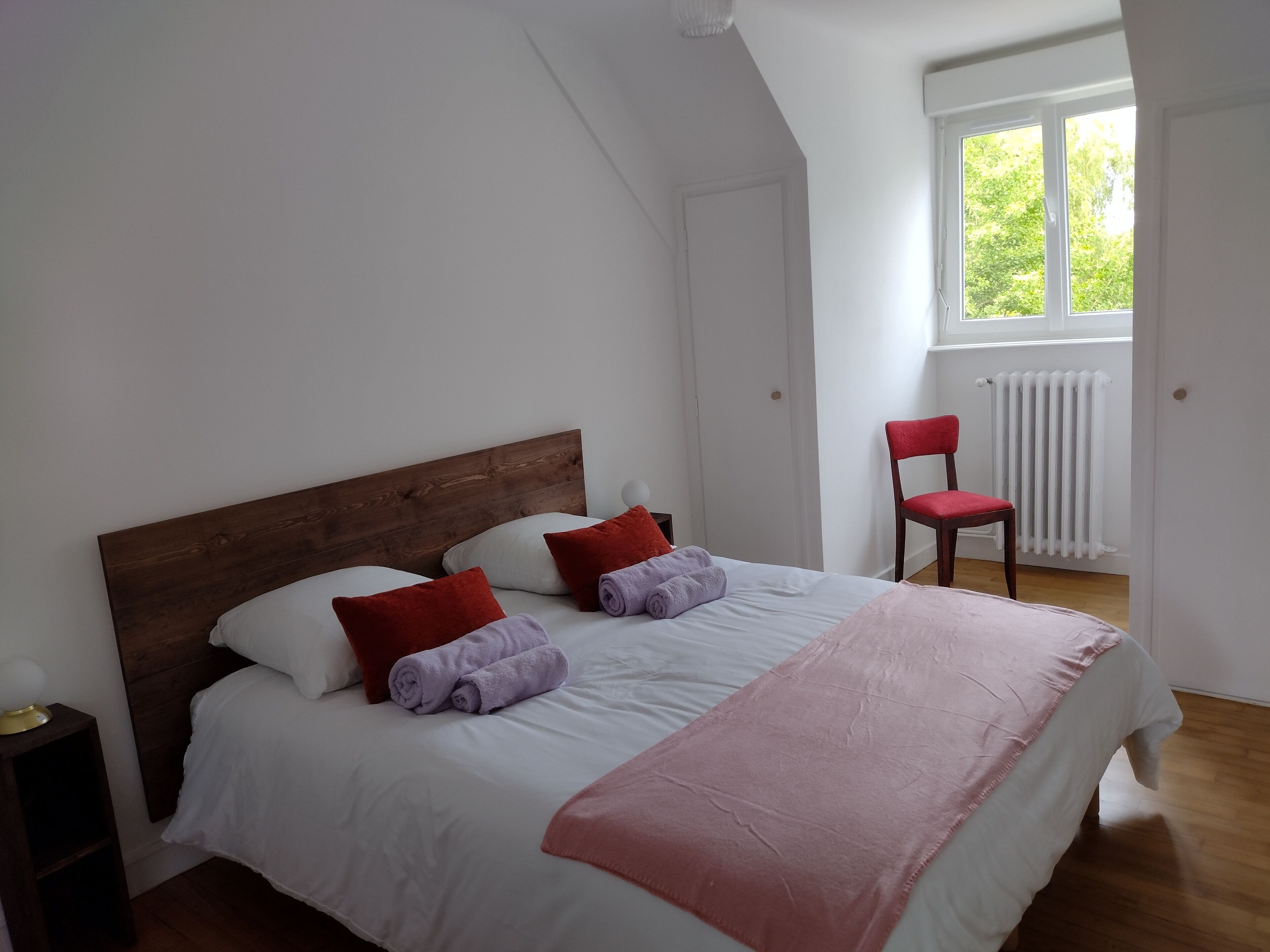 Ein ordentlich bezogenes Bett mit weißer und rosa Bettwäsche, einem Holz-Kopfteil und roten Kissen.