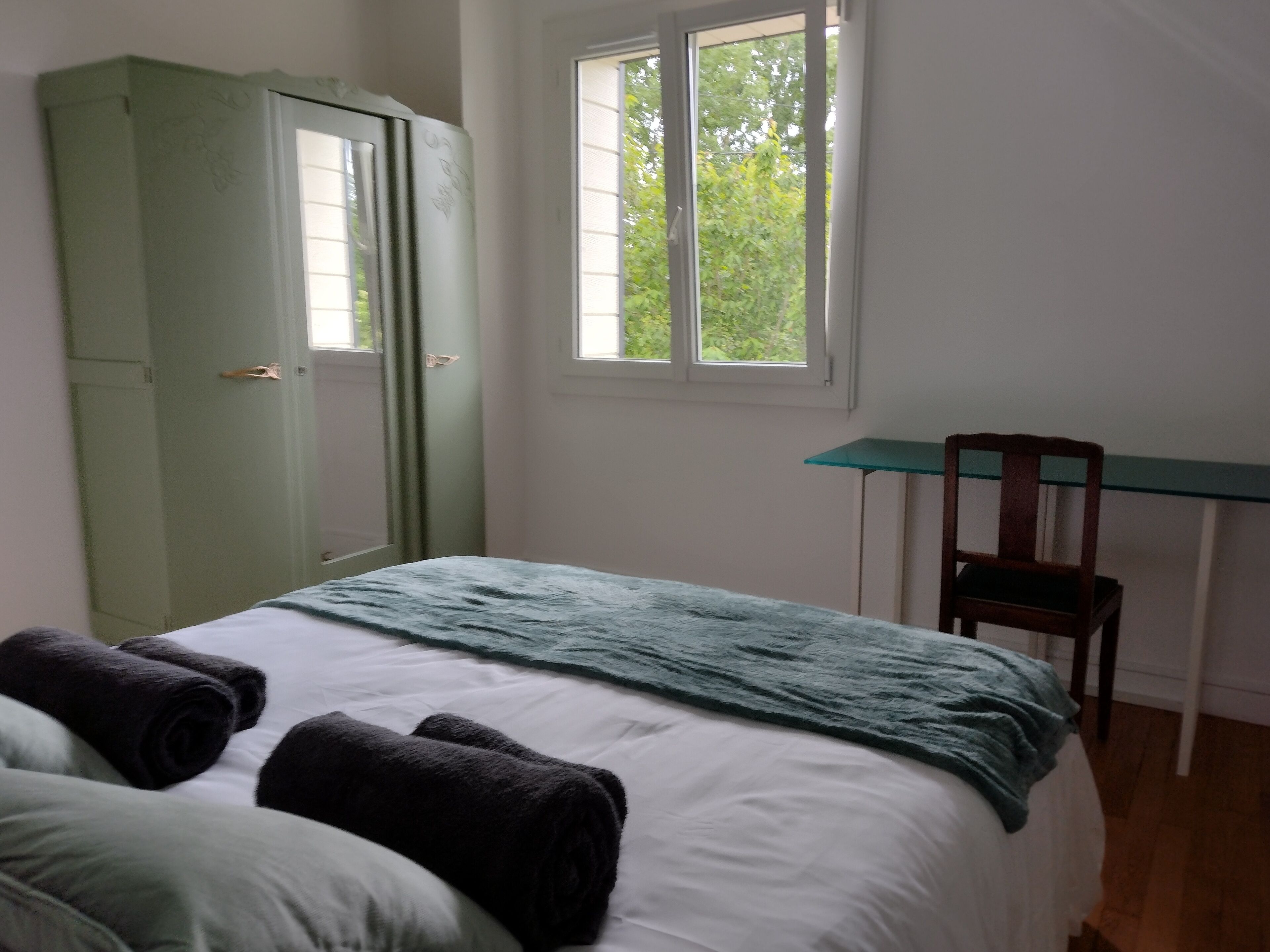 Une chambre à coucher avec un lit, une armoire verte, un bureau avec une chaise et une fenêtre donnant sur des arbres.
