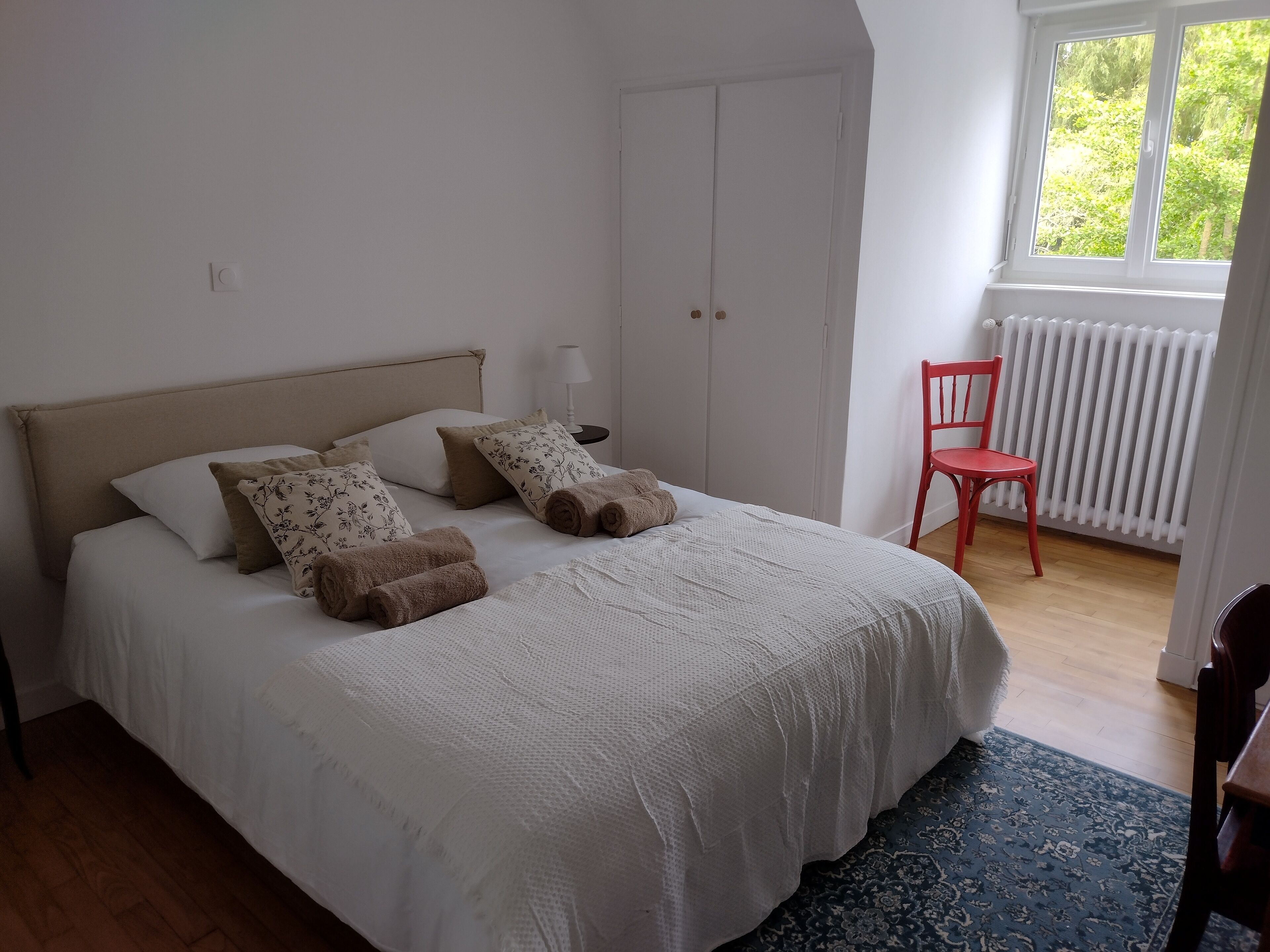 Un dormitorio con cama, almohadas, una silla, un radiador y una ventana.