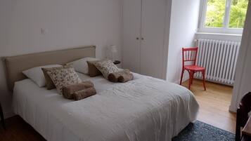 Un dormitorio con cama, almohadas, una silla, un radiador y una ventana.