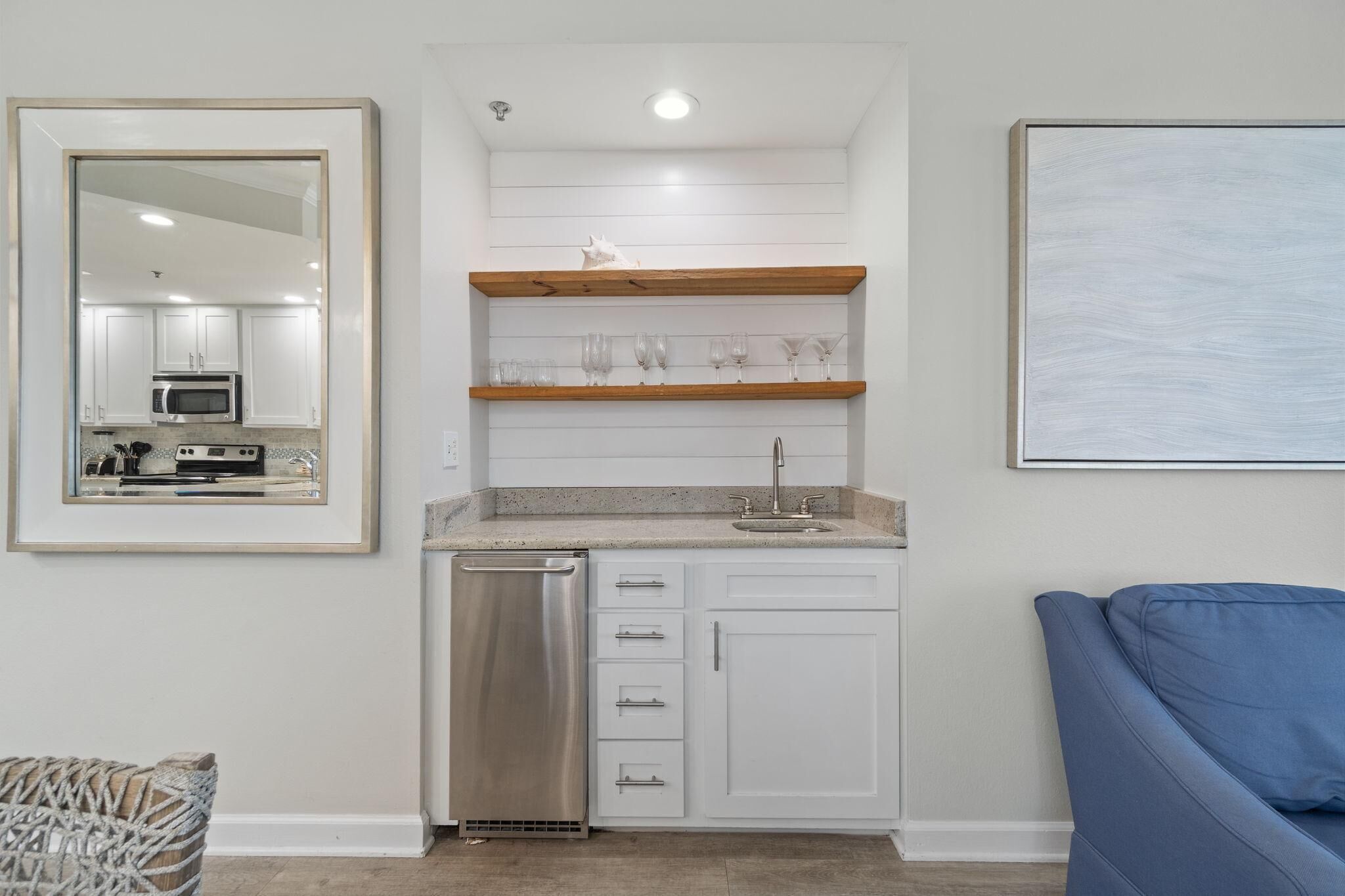 A compact kitchen with white cabinetry, a dishwasher, and a countertop with a sink. There are wooden shelves above the countertop, and a framed mirror on the left. A blue couch is visible on the right.