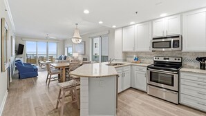A modern kitchen with white cabinets, a large island, stainless steel appliances, and a dining area with blue chairs. - Enclave 607 (Orange Beach)