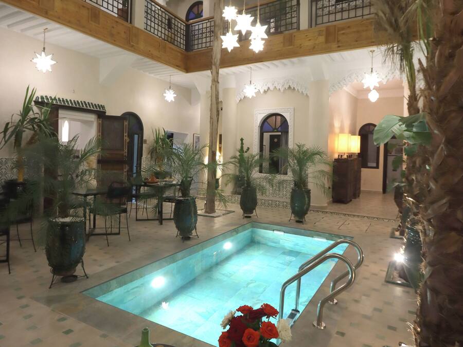 A multi-level indoor pool area with a central pool, surrounded by tables and chairs, and a staircase leading to an upper level.