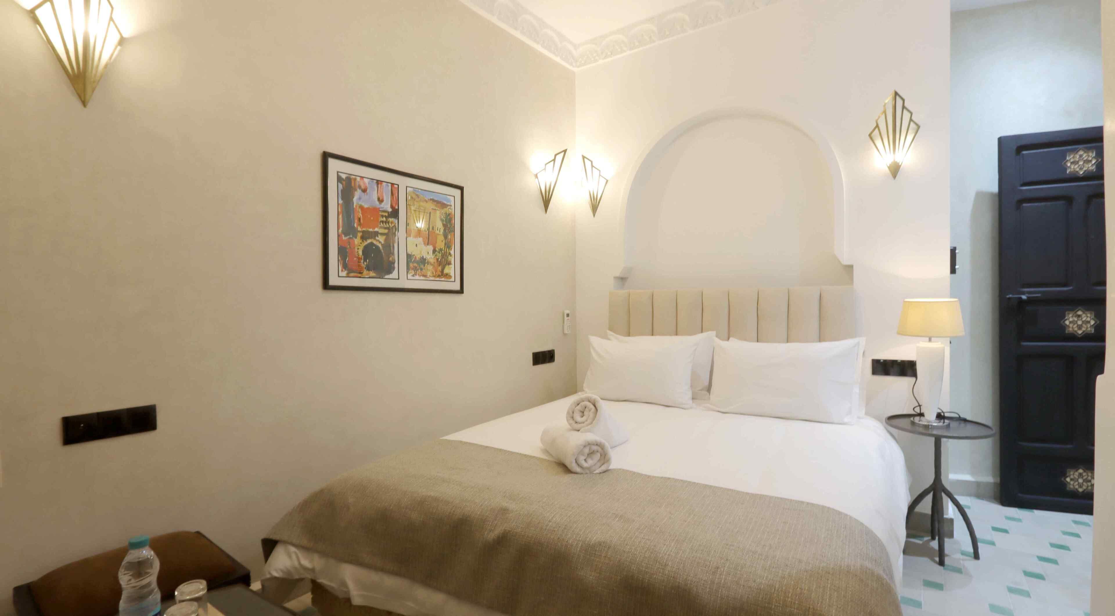 A hotel room with a bed, a nightstand, a lamp, a door, and a wall-mounted picture.