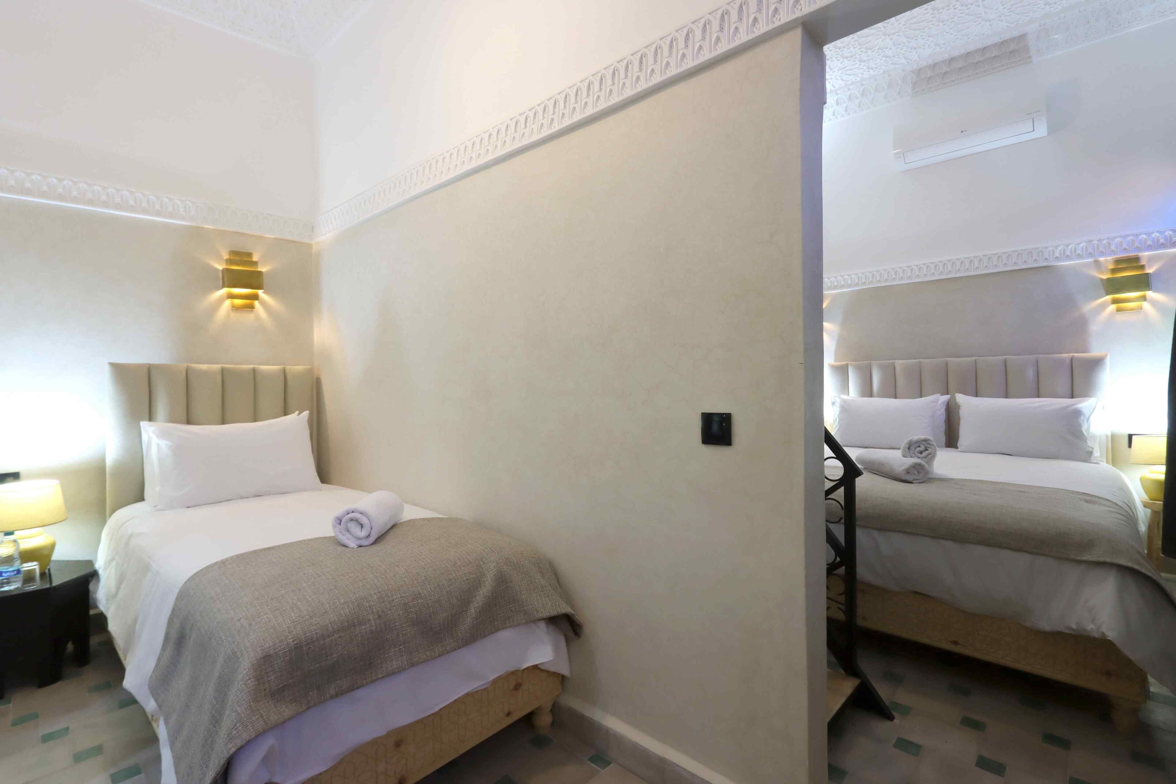A hotel room with two beds, a nightstand with a lamp, and a mirror reflecting the room.