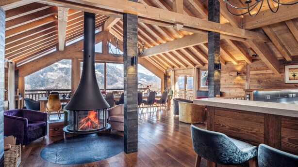 A spacious wooden interior with a central fireplace, large windows, and a modern kitchen. - Luxury Catered Ski Chalet Iona in Meribel, France LMRB-9 (Meribel)