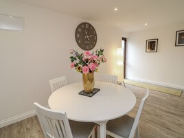 A dining area with a round white table, four white chairs, a clock on the wall, and a vase with flowers on the table.