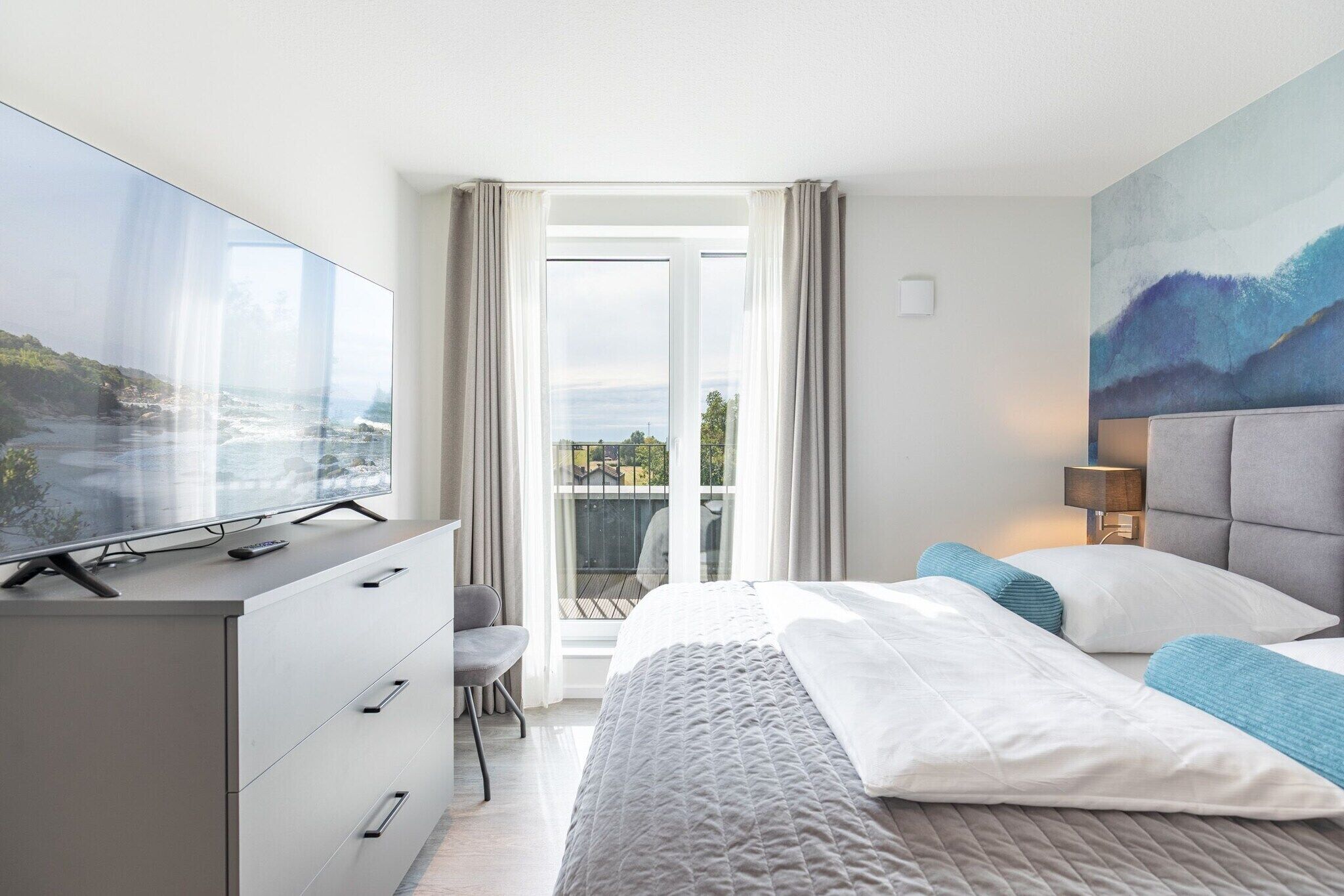 A modern hotel room with a large bed, a TV on a dresser, a balcony with a view, and a painting on the wall.