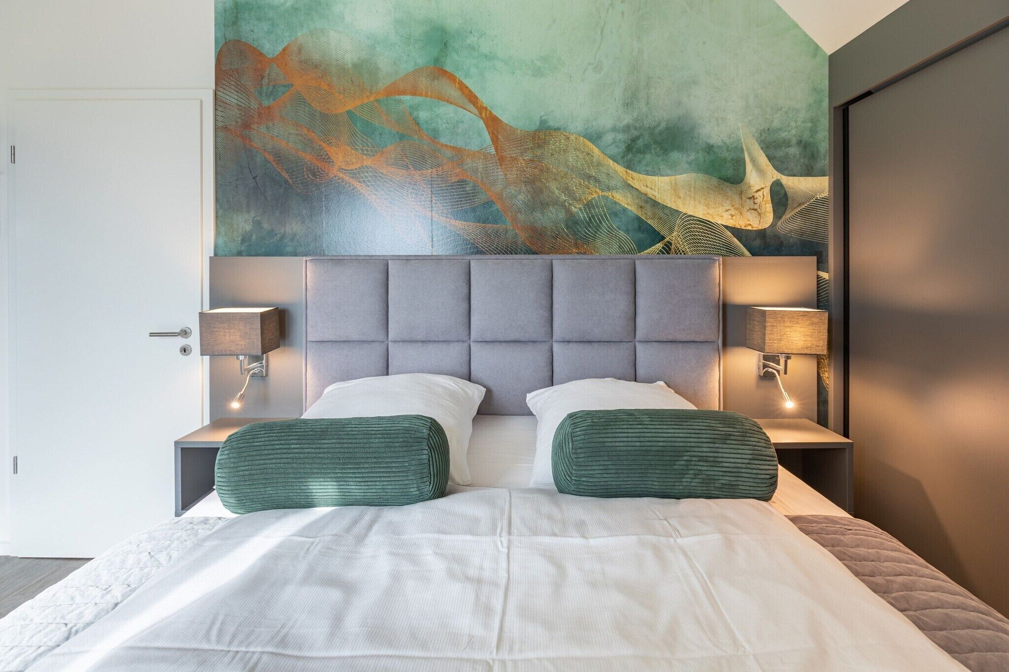 Una moderna camera d'albergo con un letto grande, due comodini con lampade, una testiera imbottita grigia e un grande quadro astratto appeso al muro.