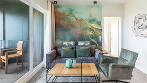Ein modernes Wohnzimmer mit einer Couch, einem Couchtisch und einem Sessel. An der Wand hängt ein großes abstraktes Gemälde, und eine Tür führt in einen weiteren Raum.
