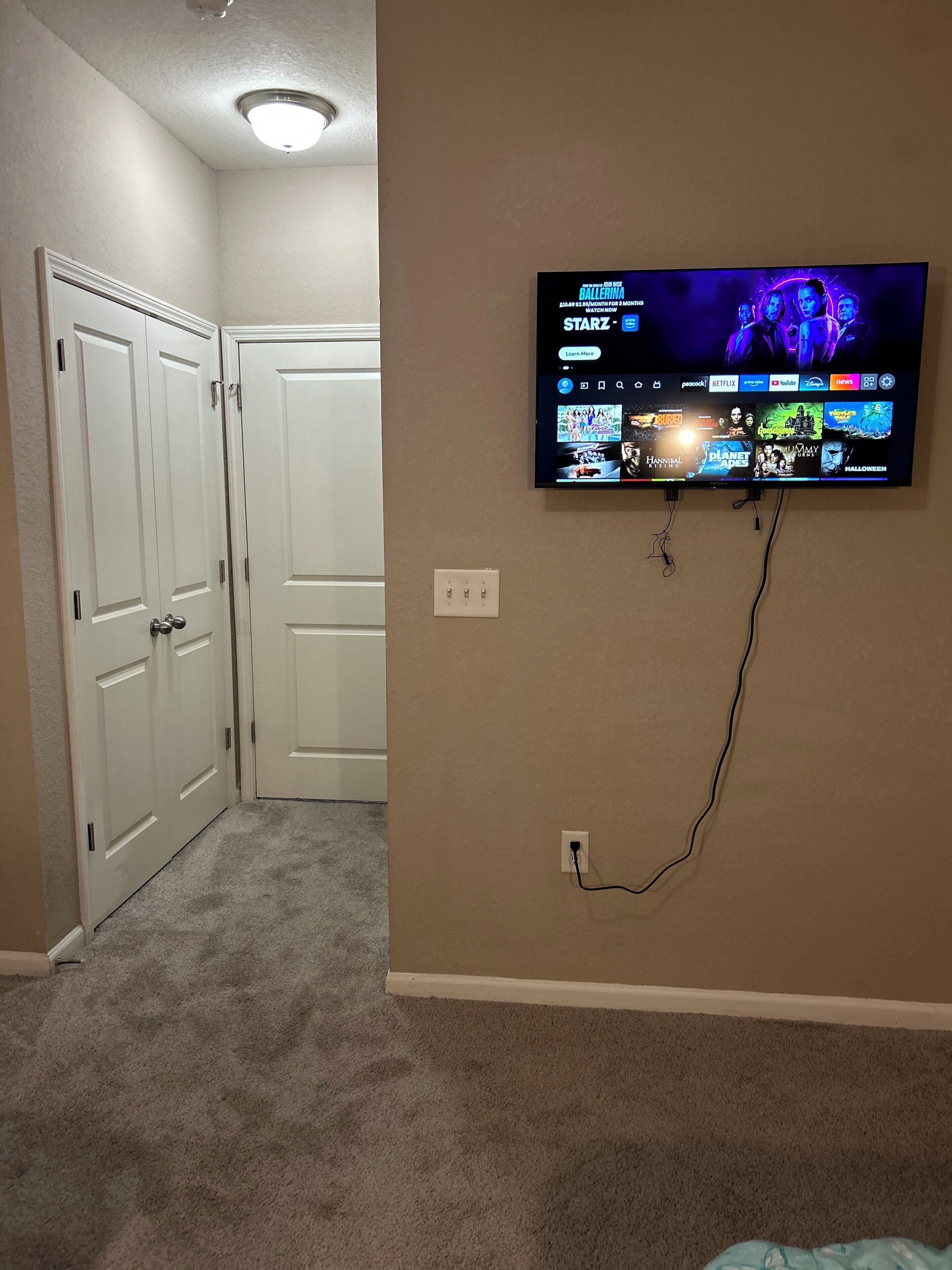 Une pièce dotée d’un téléviseur à écran plat fixé au mur, qui diffuse différents services de streaming. Les portes sont blanches et le sol est recouvert d’une moquette.