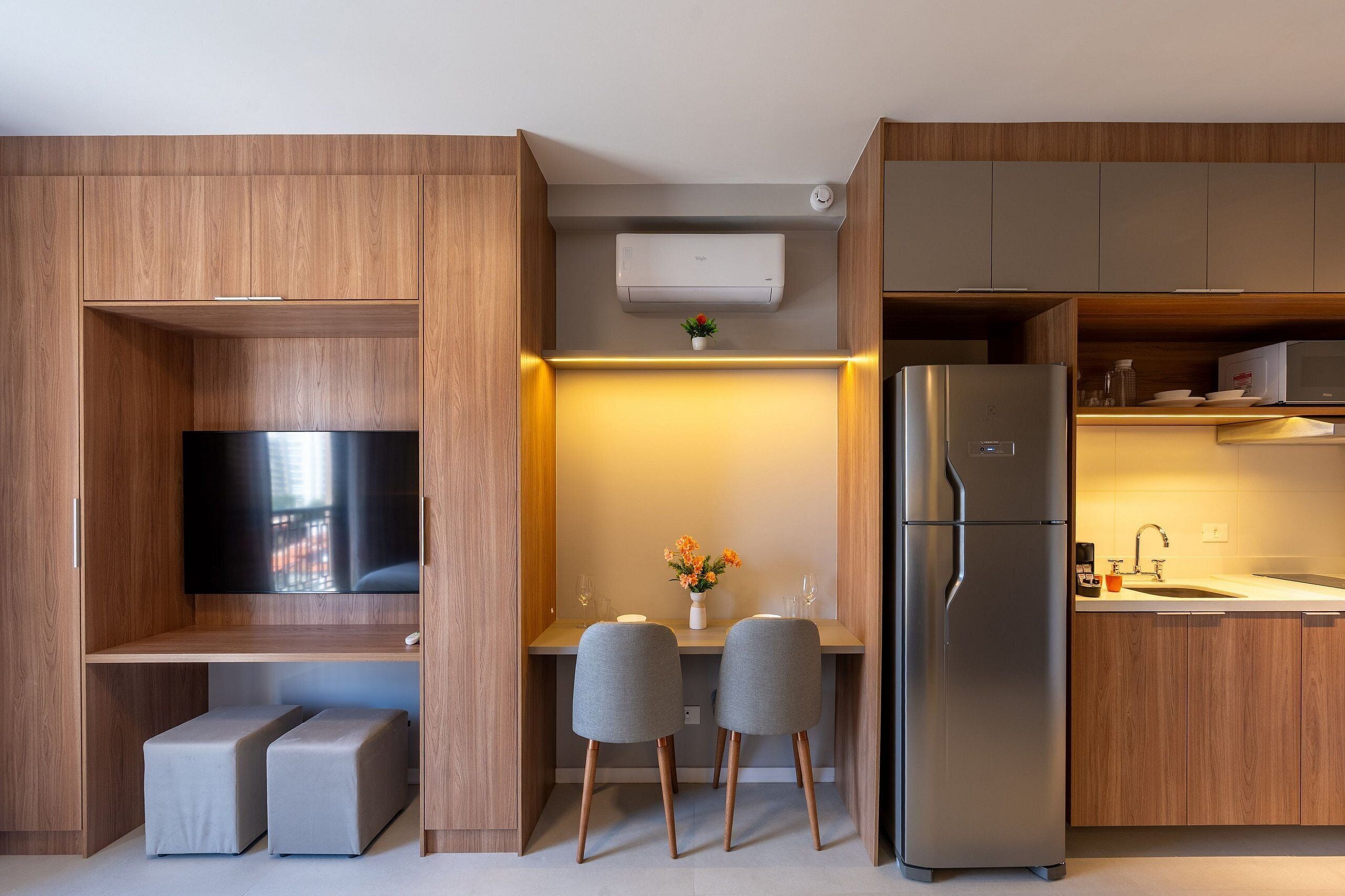 A modern kitchen with wooden cabinetry, a built-in TV, a refrigerator, and a small dining area with two chairs and a vase of flowers.