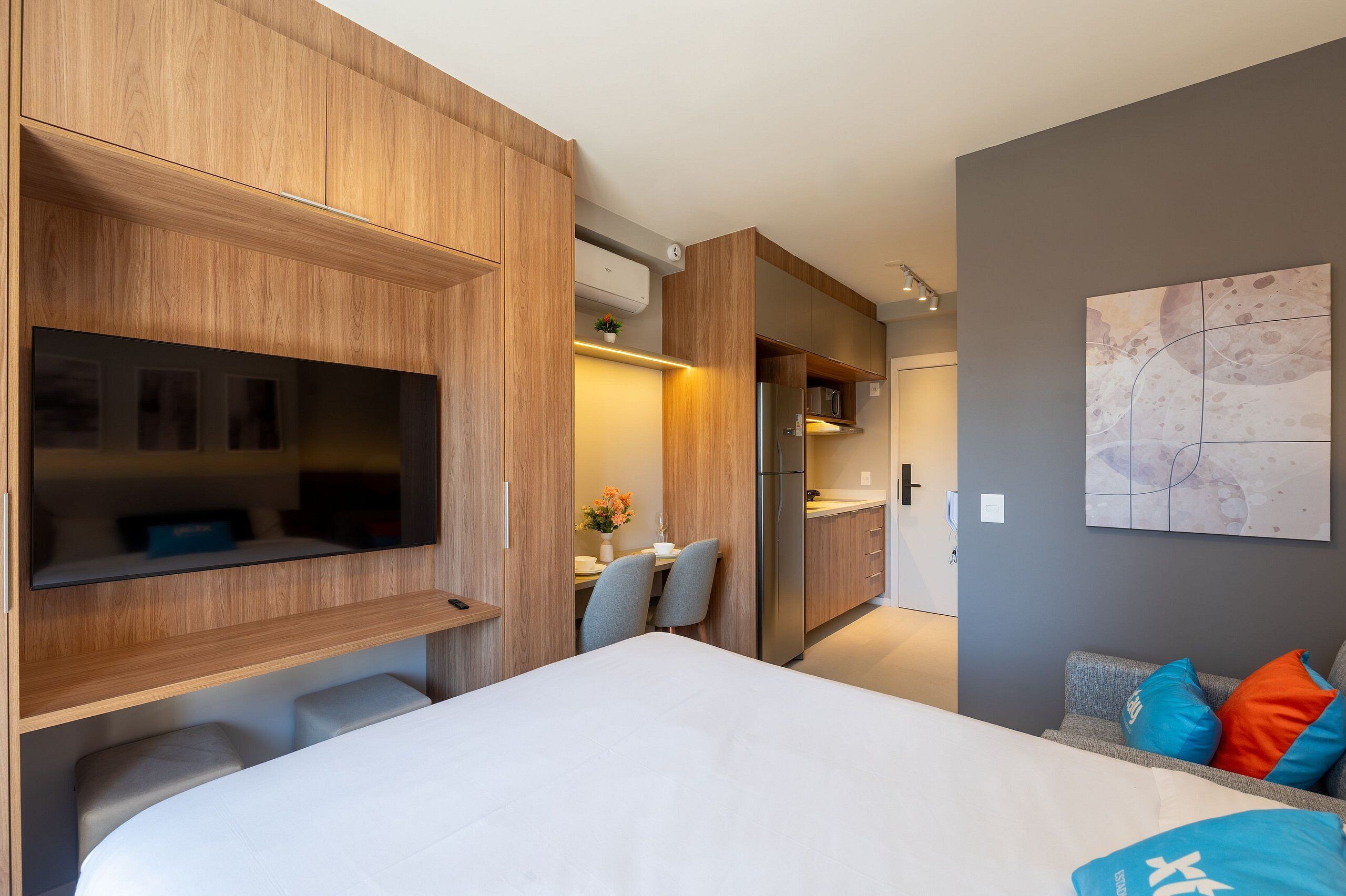 A modern hotel room with a large bed, a dining area, a kitchenette, and a living area with a TV and artwork on the wall.
