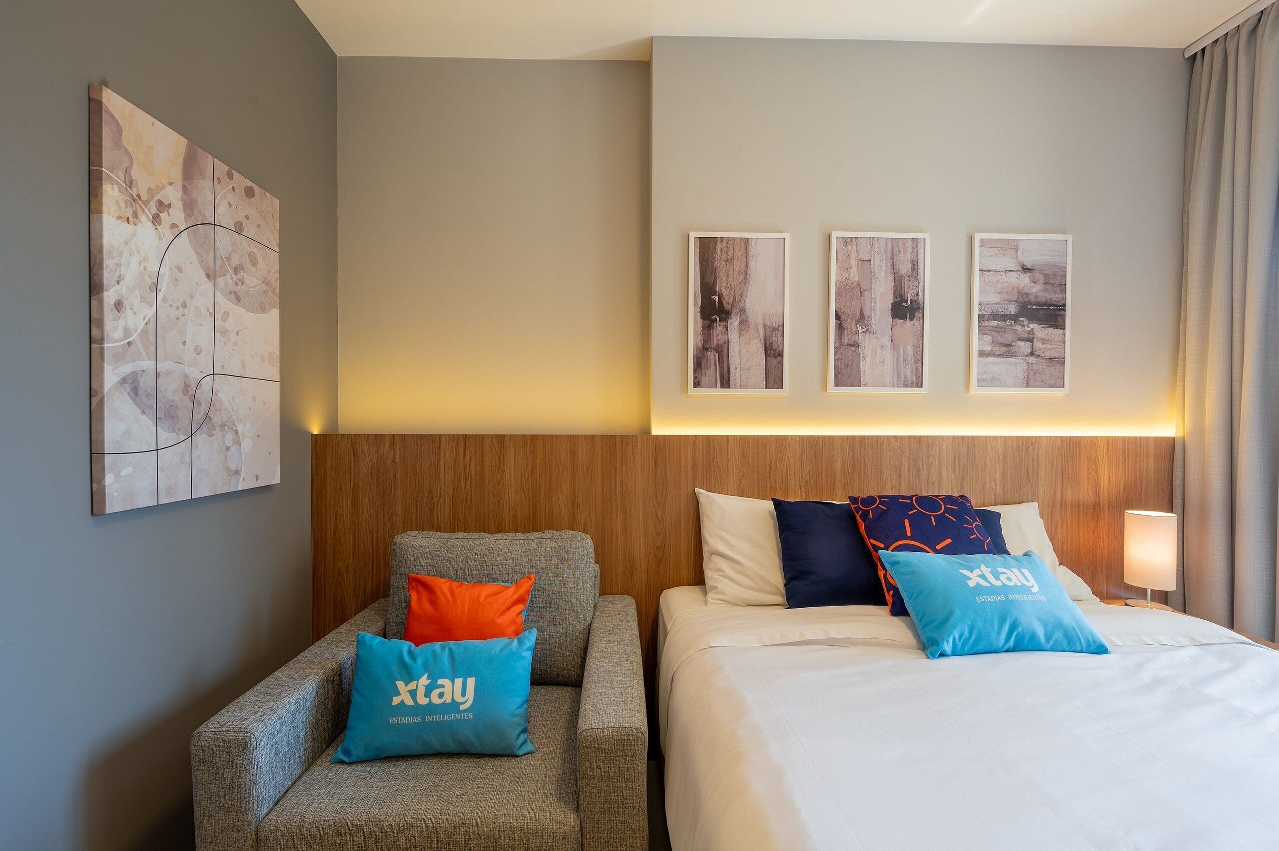 Habitación de hotel con cabecera de madera, sofá gris y cama con almohadas azules y anaranjadas.