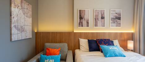 Habitación de hotel con cabecera de madera, sofá gris y cama con almohadas azules y anaranjadas.