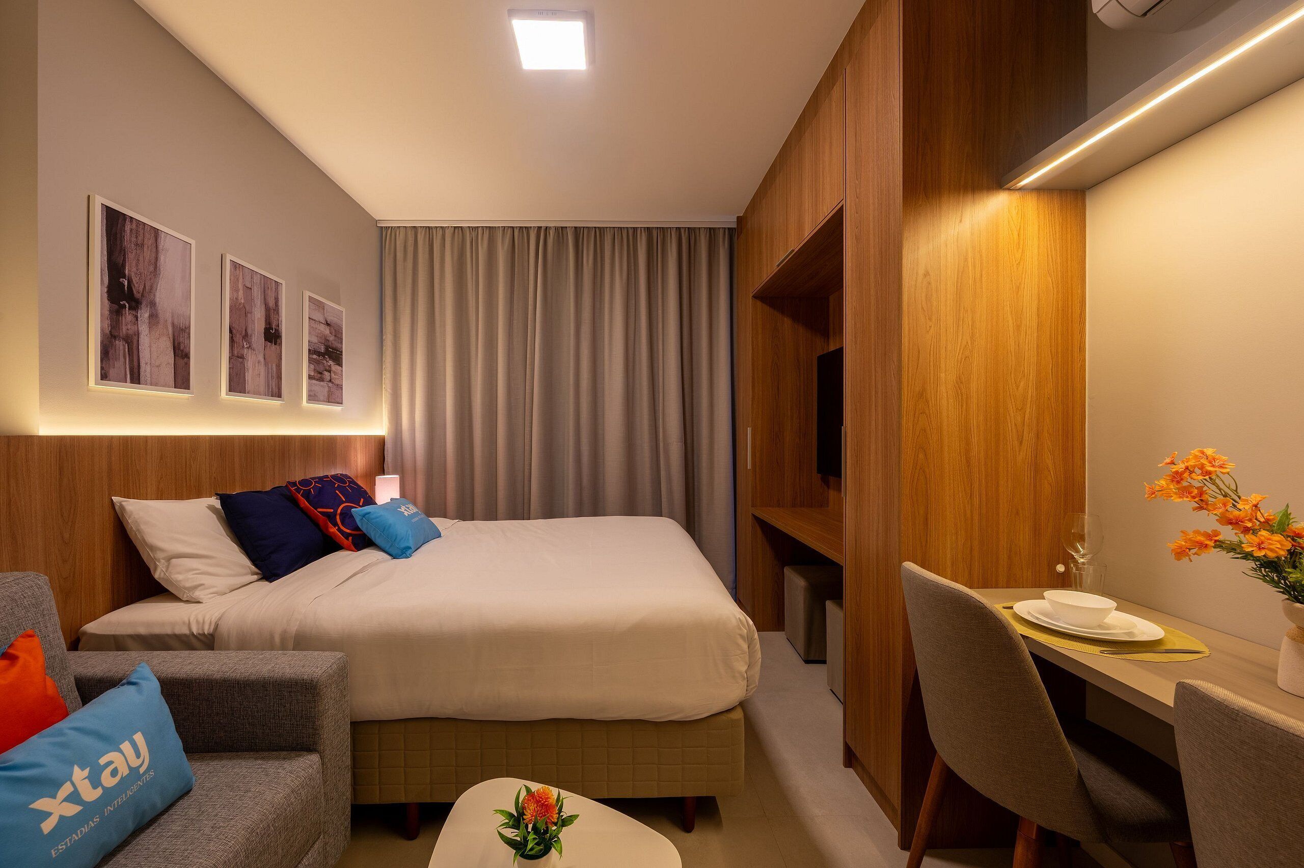 Ein Hotelzimmer mit einem Bett, einem Schreibtisch und einem Stuhl. An der Wand hängen drei gerahmte Bilder, ein Vorhang und auf dem Schreibtisch steht eine Blumenvase.